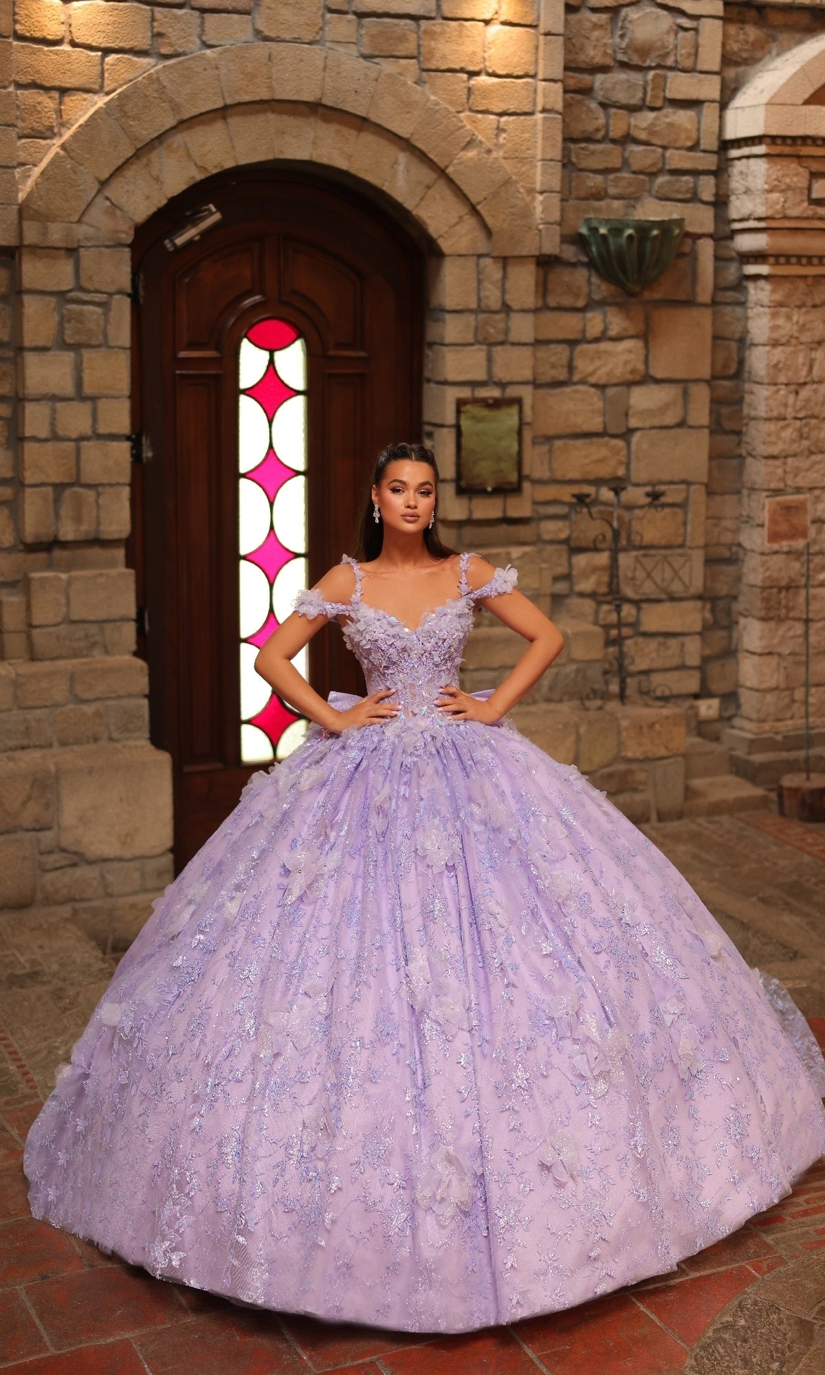 A woman stands indoors before a wooden door, wearing the Amarra 54243 off-shoulder quinceanera dress—an ornate lavender gown with a full skirt and 3D floral appliques.